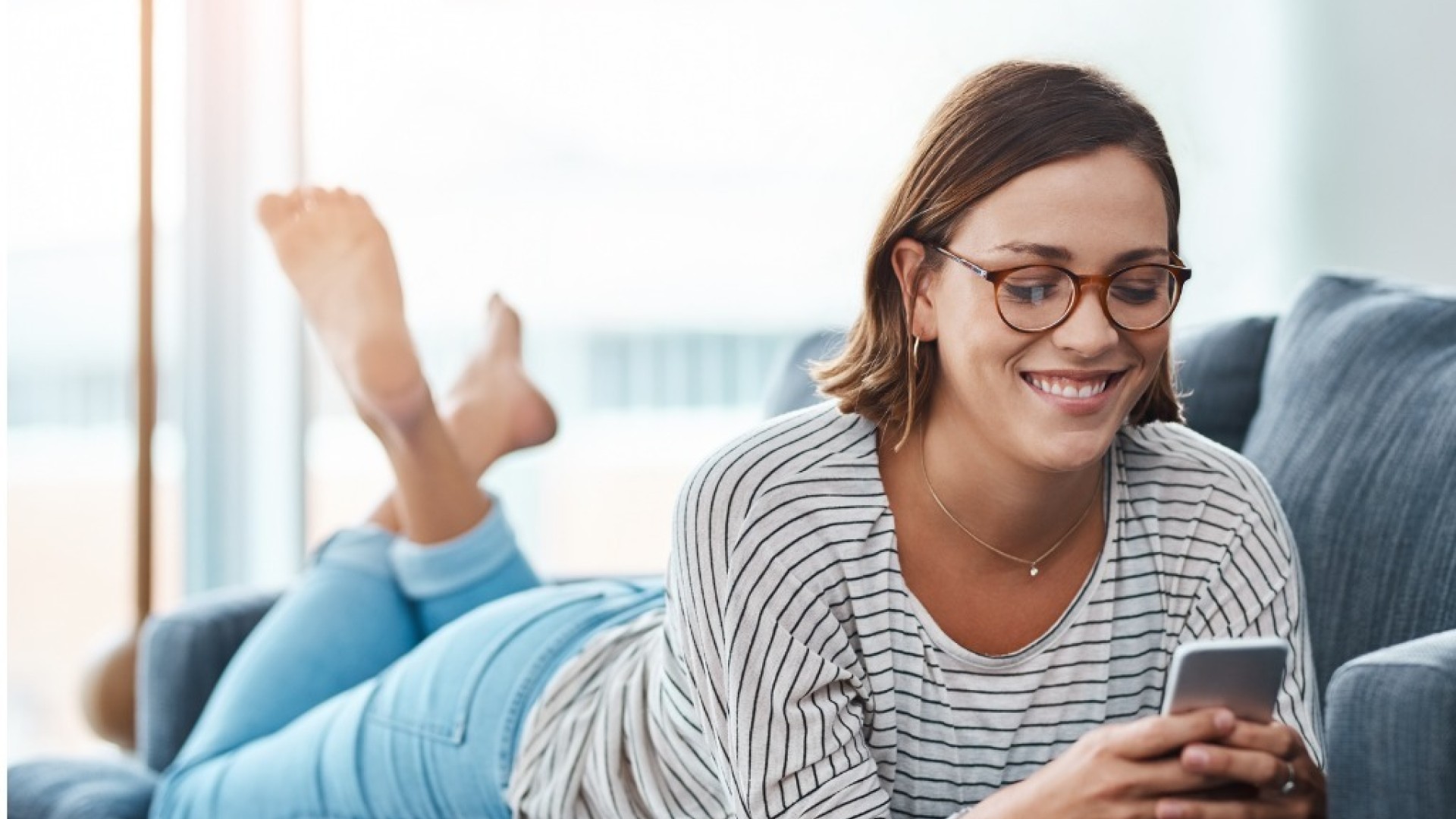 Vrouw ligt op de bank en glimlacht terwijl ze naar haar telefoon kijkt.