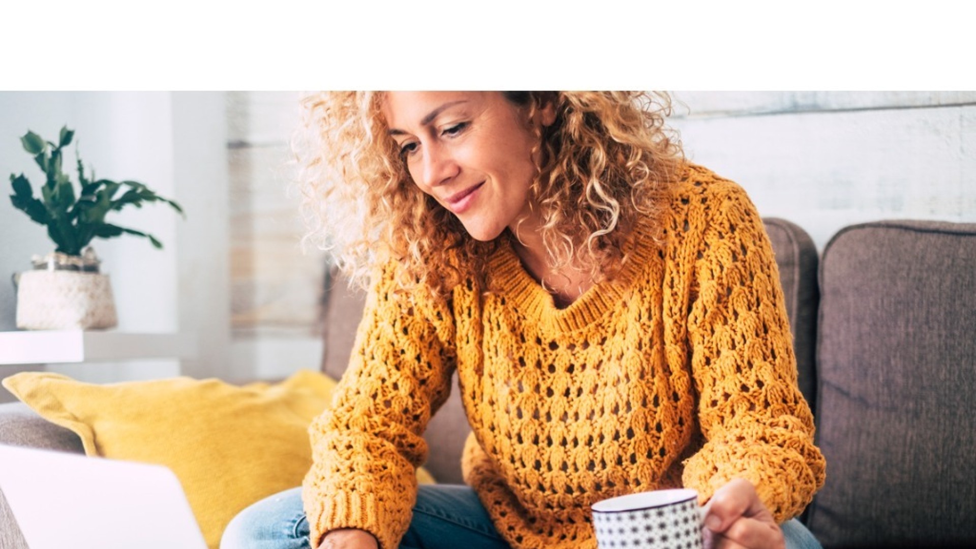 Vrouw zit op de bank met een laptop en een kopje koffie.