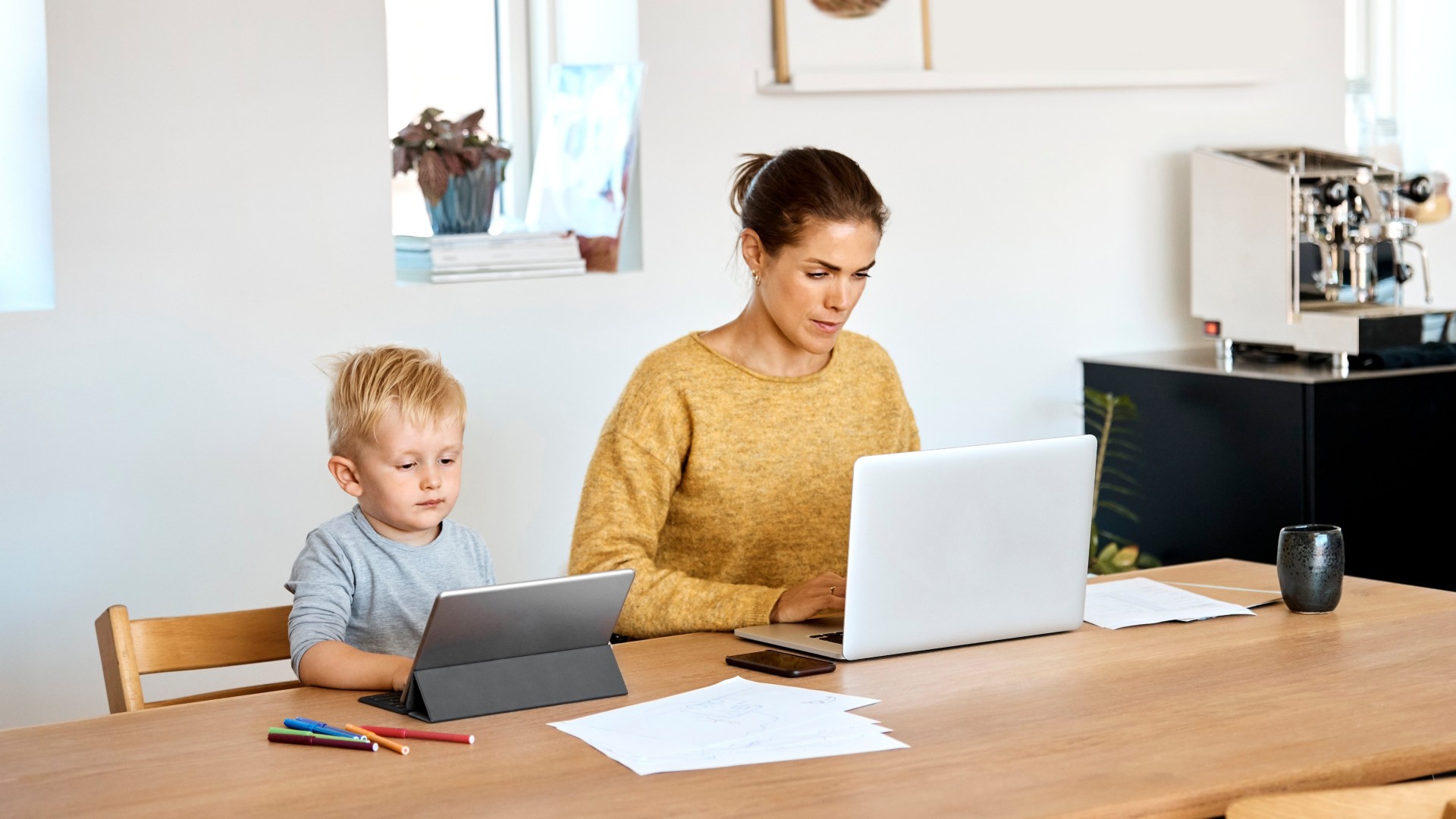 Vrouw werkt aan een laptop terwijl een kind naast haar op een tablet kijkt.