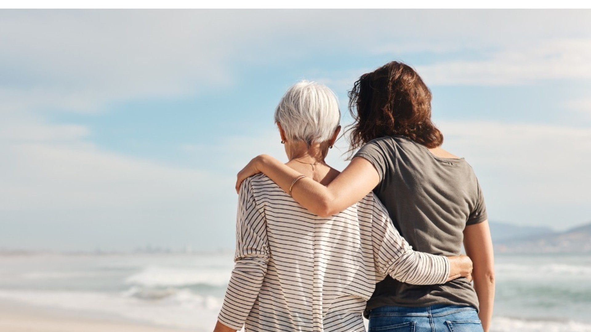 Vrouw omhelst oudere vrouw terwijl ze samen naar de zee kijken.